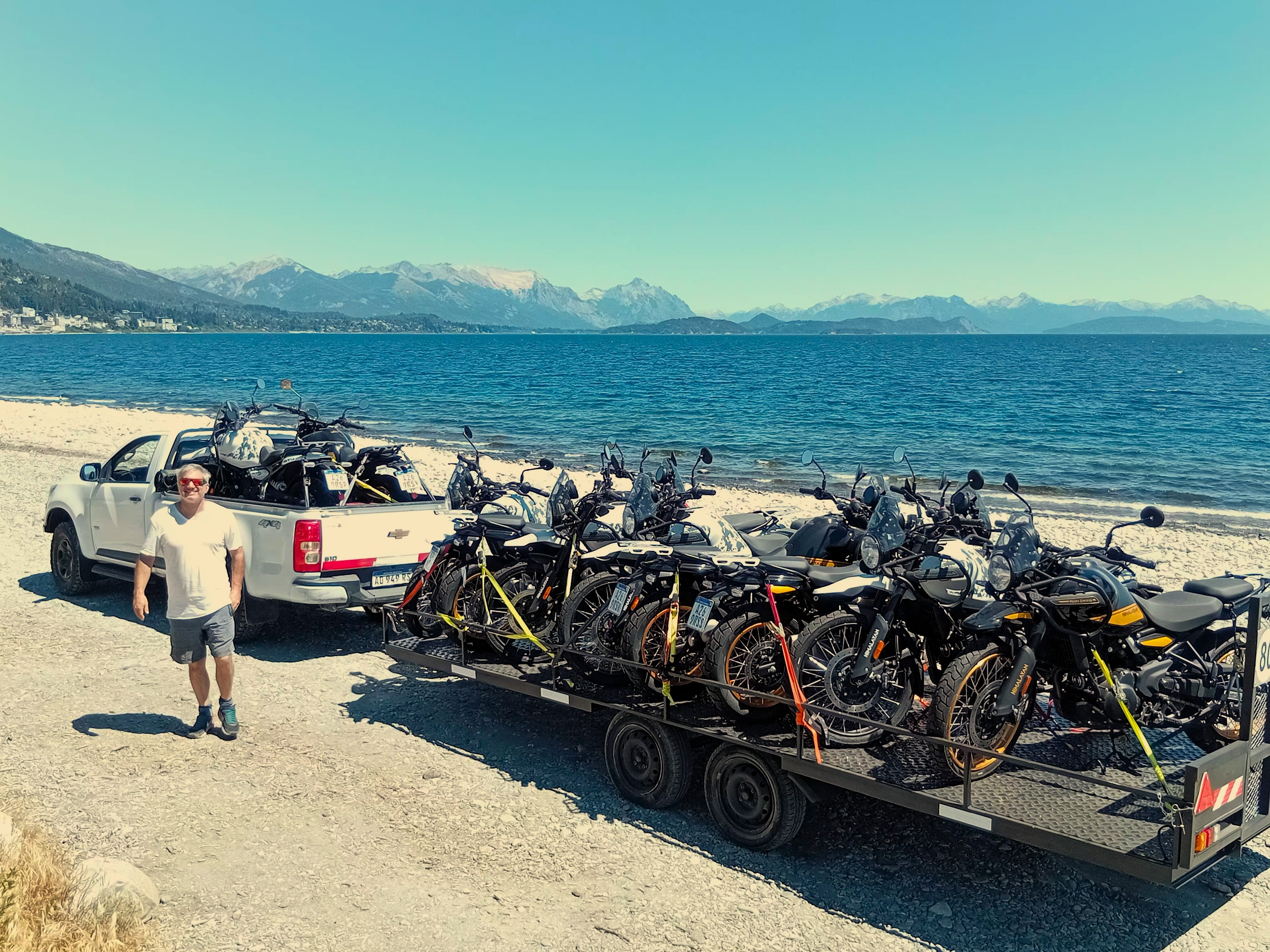 A man standing beside a trailer loaded with multiple adventure motorcycles on a sandy beach, with a blue lake, mountains, and a clear sky in the background.