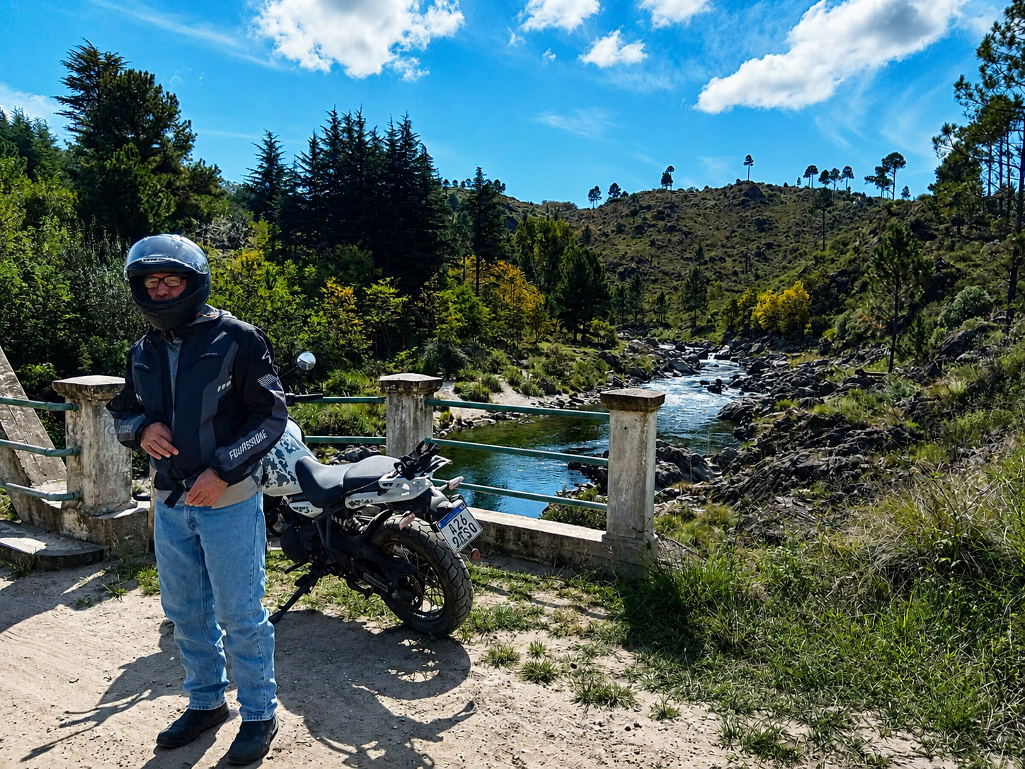Motorcyclist standing beside a BMW GS motorcycle next to a river, surrounded by mountainous landscapes in Argentina.